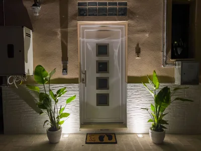 Ingresso esterno illuminato di sera con porta bianca, piante ornamentali e zerbino di benvenuto