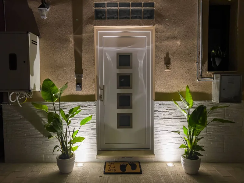Ingresso esterno illuminato di sera con porta bianca, piante ornamentali e zerbino di benvenuto